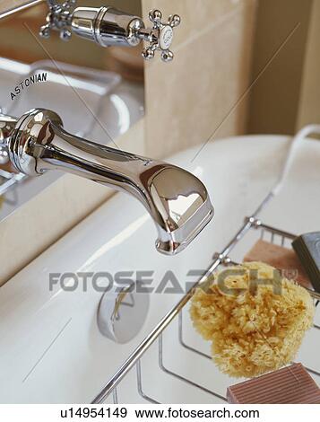 Stock Photo - Close-up of stainless steel tap above bath with sponge on metal bath rack. Fotosearch