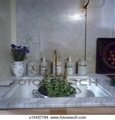 Picture - Close-up of tap with water running into vegetables in colander in marble sink in front of marble splashback. Fotosearch