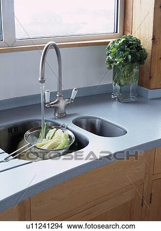 Picture - Close-up of water pouring from stainless steel mixer tap into vegetables in sieve in double underset sink in grey worktop. Fotosearch