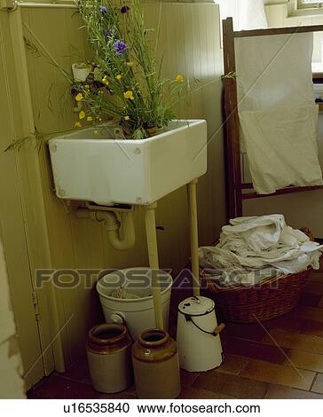 Stock Image - Corner of old-fashioned laundry room with white ceramic sink above earthenware storage jars and basket of washing. Fotosearch
