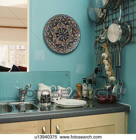Stock Photography - Corner of turquoise kitchen with ceramic plate on wall beside pans on metal rank above stainless steel sink. Fotosearch