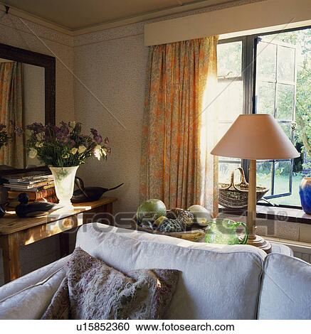 Stock Image - Country livingroom with lamp with peach lampshade on small table behind sofa. Fotosearch