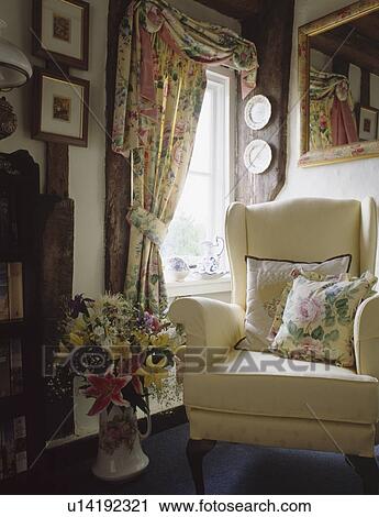 Stock Image - Cream armchair and floral curtains in cottage living room. Fotosearch