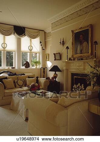 Picture - Cream blind and draped curtains in neutral living room with picture above fireplace and cream sofas. Fotosearch