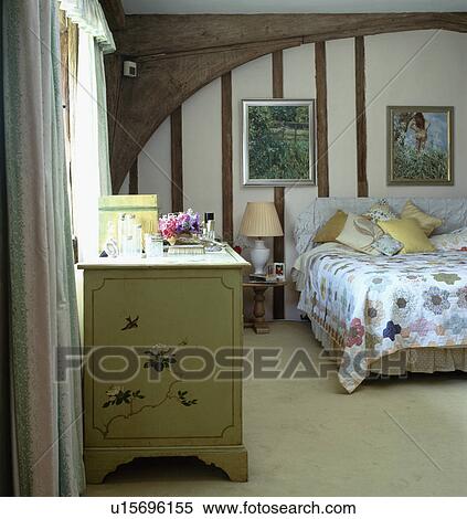 Stock Photography - Cream carpet and chest of drawers in country bedroom with beamed walls and patchwork quilt on bed . Fotosearch