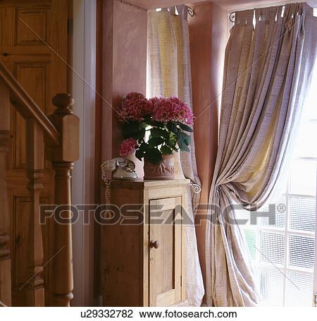 Stock Image - Cream curtain on window beside small pine cabinet with telephone and pink plant in hall. Fotosearch