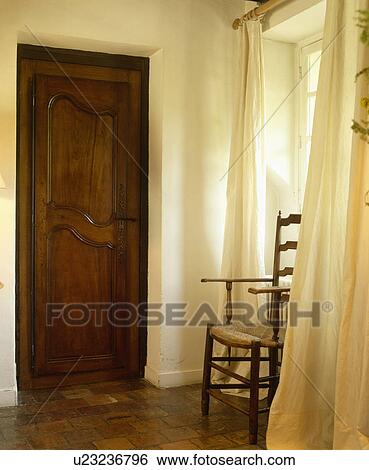 Stock Photograph - Cream curtains at window in hall with rush-seated chair and old mahogany door. Fotosearch