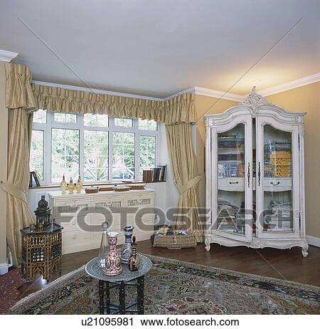 Cream silk curtains and glass-fronted white painted cupboard in bedroom with Moroccan jugs and tables View Large Photo Image Stock Image - Cream silk curtains and glass-fronted white painted cupboard in bedroom with Moroccan jugs and tables. Fotosearch