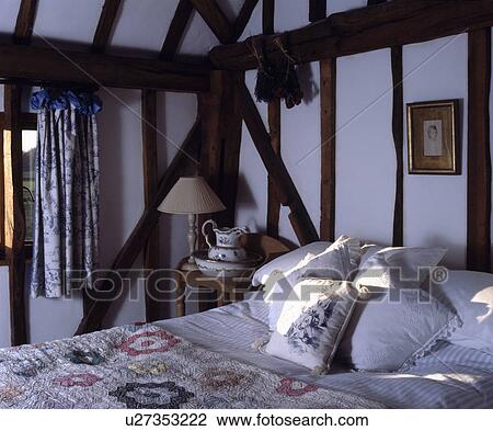 Stock Image - Cushions on bed in small cottage bedroom with exposed wall beams. Fotosearch
