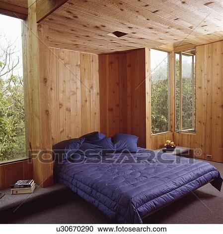 Stock Image - Dark blue pillows and quilt on bed in log cabin bedroom with wooden walls and ceiling. Fotosearch