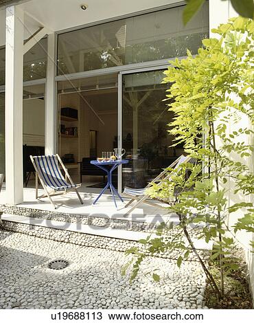 Deckchair and table on pebble paved patio in front of modern house with glass doors View Large Photo Image Stock Image - Deckchair and table on pebble paved patio in front of modern house with glass doors. Fotosearch