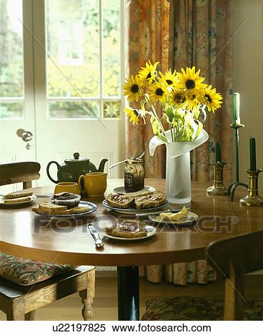 Stock Photography - Dining room with yellow sunflowers in glass vase on circular wood table set for breakfast with teapot and plates of toast. Fotosearch