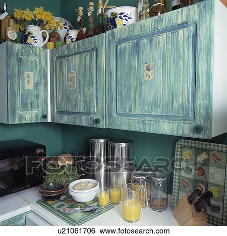 Stock Photograph - Distressed blue ragged paint effect fitted cupboards above metal cannisters and glasses of fruit juice on kitchen worktop. Fotosearch