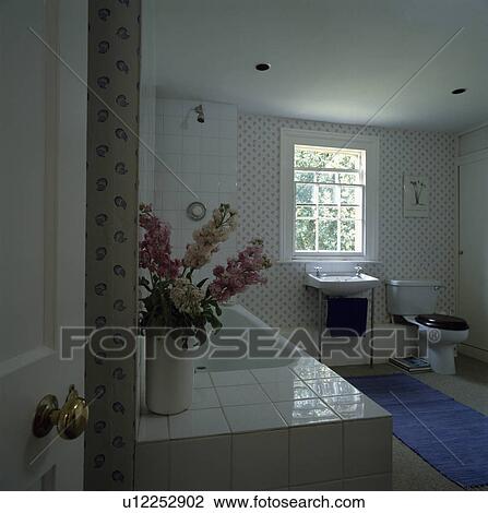 Stock Image - Door open to traditional bathroom with vase of pink stocks on bath with white tiled surround . Fotosearch