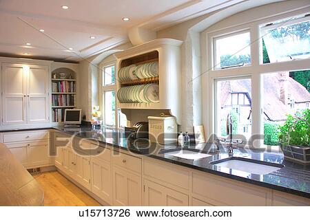 Stock Photograph - Double ceramic sinks underset into black granite worktops below window in white contemporary kitchen. Fotosearch