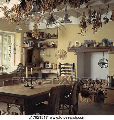 Stock Photo - Dried herbs and hops on beams above antique pine table and chairs in pastel yellow country kitchen. Fotosearch