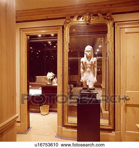 Stock Image - Egyptian sculpture on plinth in front of tall gilt mirror on landing with open door to bathroom. Fotosearch