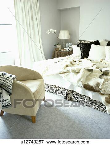 Faux Fur Throws And Cushions On Bed In Modern Grey Bedroom With Cream Suede Upholstered Chair And Grey Carpet Stock Photo