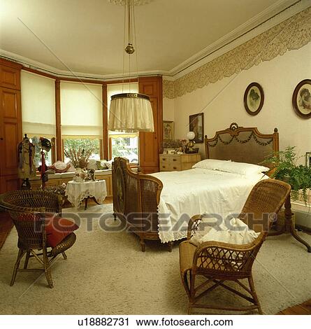Stock Image - French Bergere bed and cane chairs in bedroom with cream blinds and carpet. Fotosearch
