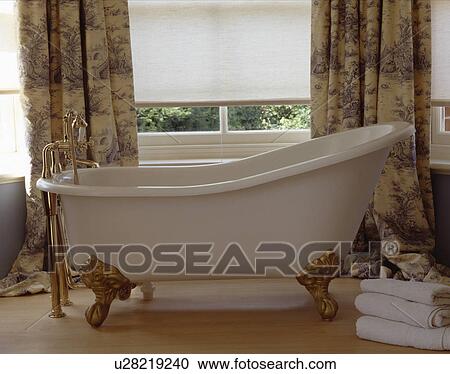 Stock Image - Gilt claw feet on roll top bath in front of window with white blind and Toile de Jouy curtains. Fotosearch