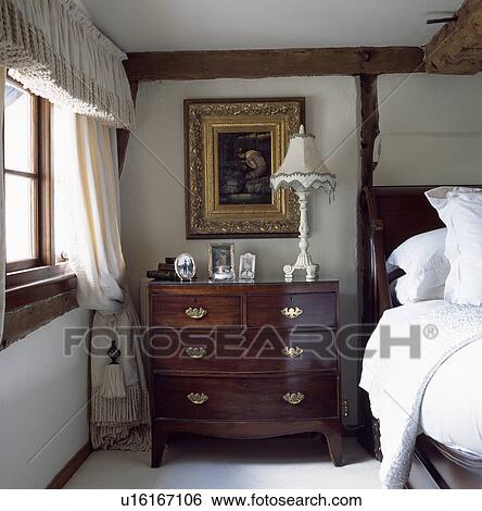 Gilt-framed picture above antique chest-of-drawers with lamp in cottage bedroom with tasselled tieback on cream curtains View Large Photo Image Stock Photograph - Gilt-framed picture above antique chest-of-drawers with lamp in cottage bedroom with tasselled tieback on cream curtains. Fotosearch