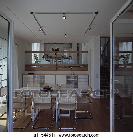 Glass doors open to cane and chrome chairs and white table in openplan white eighties dining room View Large Photo Image Stock Image - Glass doors open to cane and chrome chairs and white table in openplan white eighties dining room. Fotosearch
