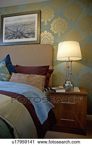 Stock Image - Gold-patterned green wallpaper in modern bedroom with beige upholstered headboard and lighted modern lamp on small bedside cabinet. Fotosearch