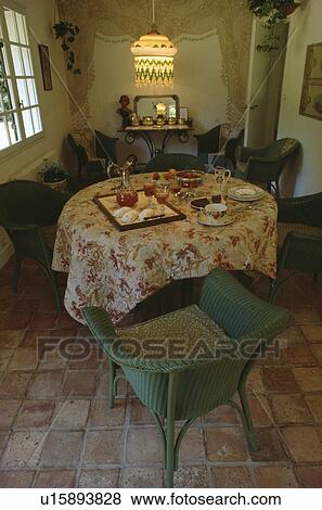 Stock Photo - Green Lloyd Loom chair beside circular table with floral cloth and place settings in dining room with beaded lampshade. Fotosearch