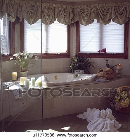 Stock Photograph - Grey festoon blinds at windows above bath and white towels on floor of modern bathroom. Fotosearch