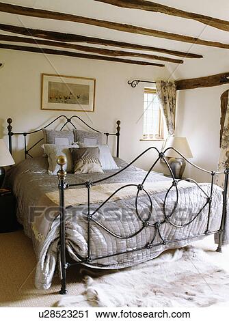 Stock Image - Grey silk quilt and cushions on modern wrought iron bed in cream cottage bedroom. Fotosearch