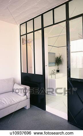 Stock Photograph - Grey sofa beside black painted and glazed door way leading to large white contemporary bathroom. Fotosearch