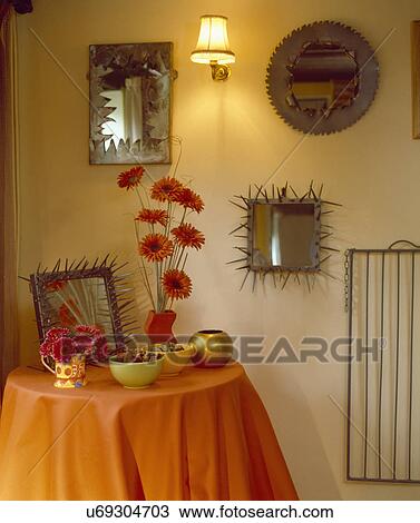 Stock Image - Handmade mirrors with beaten metal frames on wall above circular table with red gerberas on orange cloth . Fotosearch