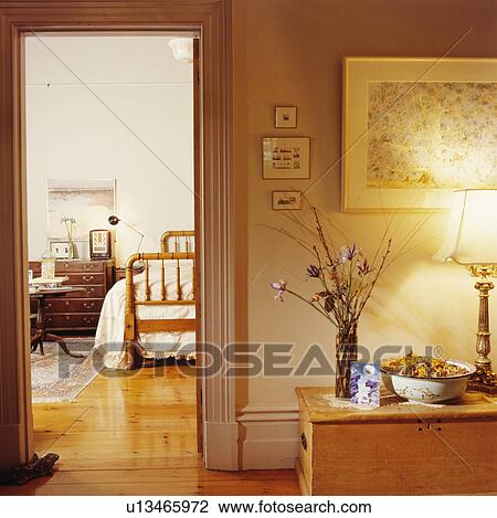 Stock Image - Landing with lighted lamp on pine chest beside open door with view of traditional bedroom with wooden floor. Fotosearch