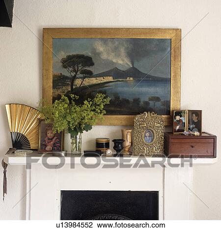 Stock Image - Landscape painting above mantelpiece with photographs and antique fan with Treen ornaments. Fotosearch