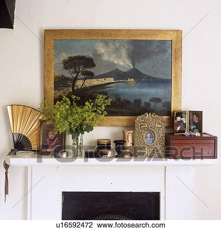 Stock Image - Landscape painting above mantelpiece with photographs and antique fan with Treen ornaments. Fotosearch