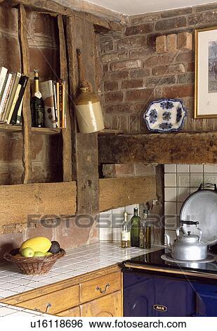 Stock Photograph - Large beams and brick wall above white tiled worktop beside blue Aga in corner of cottage kitchen. Fotosearch