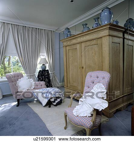 Stock Image - Large pale wood wardrobe in front of mirrored wall in bedroom with pink chaiselongue and armchair. Fotosearch