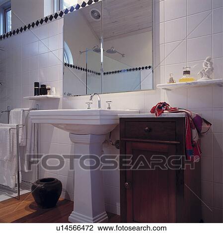Stock Image - Large rectangular mirror above white pedestal basin and small mahogany cupboard in modern white tiled bathroom. Fotosearch