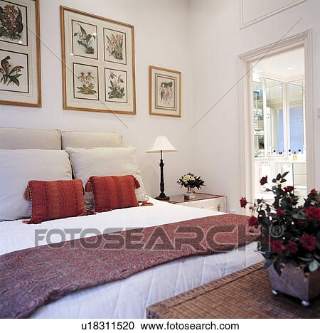 Stock Image - Large white bedroom with framed pictures on wall and red and white cushions on bed. Fotosearch