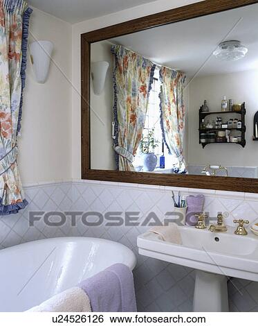 Stock Photograph - Large wooden mirror above bath and pedestal basin in small cream bathroom. Fotosearch