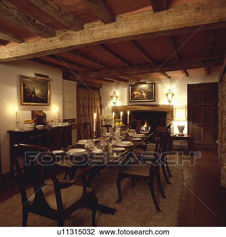 Stock Image - Lighted candles and place settings on rectangular antique oak table in country dining room with beamed ceiling. Fotosearch