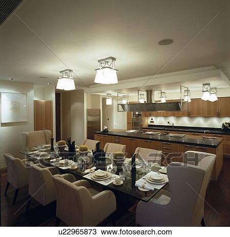 Stock Image - Lighted ceiling lights in modern kitchen dining room with place settings and beige upholstered chairs. Fotosearch