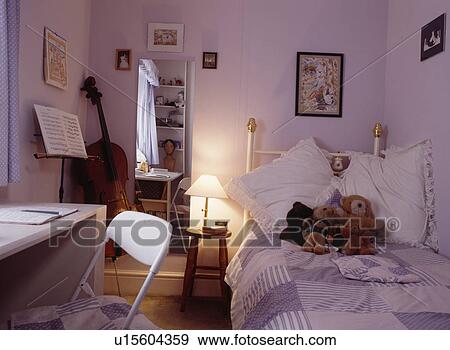 Stock Photo - Lighted lamp and mirror beside brass bed in teenager's mauve bedroom with lilac patchwork quilt and white desk and chair. Fotosearch