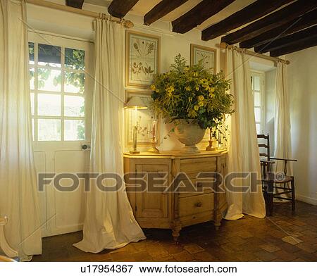 Stock Photo - Lighted lamps and flower arrangement on cupboard between windows in white cottage living room. Fotosearch