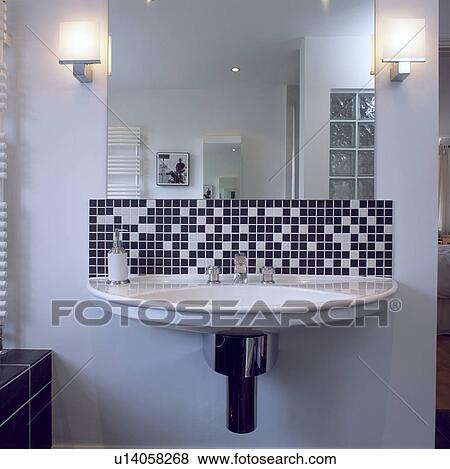 Stock Photo - Lighted lamps on either side of mirror above black+white mosaic tiles and flat white basin . Fotosearch