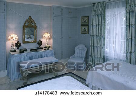 Stock Image - Lighted lamps on skirted dressingtable with upholstered stool in pastel blue bedroom with sheers and blue curtains. Fotosearch