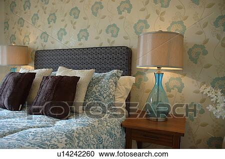 Lighted lamps with blue glass bases on either side of bed with blue and white duvet in modern bedroom with blue stylised floral patterned wallpaper View Large Photo Image Stock Image - Lighted lamps with blue glass bases on either side of bed with blue and white duvet in modern bedroom with blue stylised floral patterned wallpaper. Fotosearch