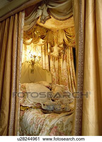 Stock Image - Lighting inside opulent gold curtains on fourposter bed with floral cushions and quilt. Fotosearch