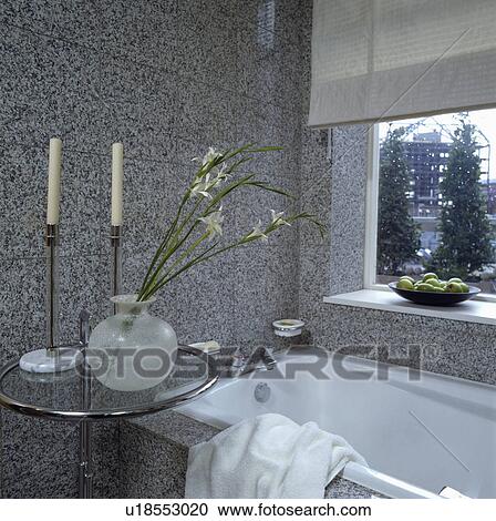 Stock Image - Lilies in spherical glass vase on chrome and glass Eileen Gray table beside bath in grey granite tiled bathroom . Fotosearch