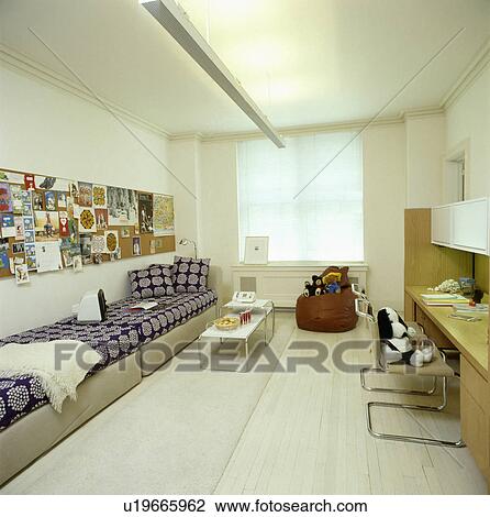 Stock Image - Long pinboard above divan bed sofas with blue+white covers in teenage beroom with white painted flooring. Fotosearch